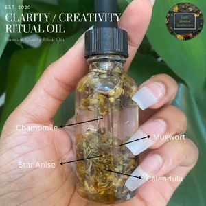 May include: A clear glass bottle filled with a blend of herbs and flowers, including chamomile, mugwort, star anise, and calendula. The bottle is labeled "Clarity/Creativity Ritual Oil" and "Premium Quality Ritual Oils".