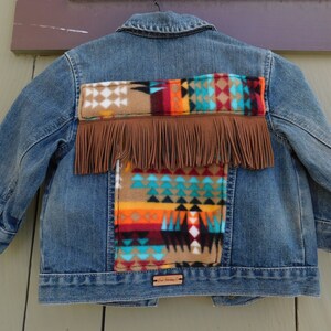May include: A blue denim jacket with a colorful geometric pattern on the back. The pattern is made of a brown, orange, red, turquoise, and black fabric with fringe trim.