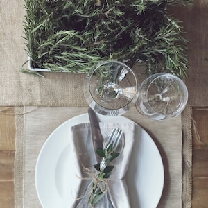 May include: A white plate with a white napkin, a fork, and a knife tied together with twine and a sprig of rosemary. Two clear wine glasses are on a beige placemat with a sprig of rosemary in the background.