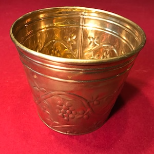 Könnte beinhalten: Ein goldfarbener Metalltopf mit einem geprägten Design aus Trauben und Reben. Der Topf ist rund und hat eine glatte, glänzende Oberfläche.