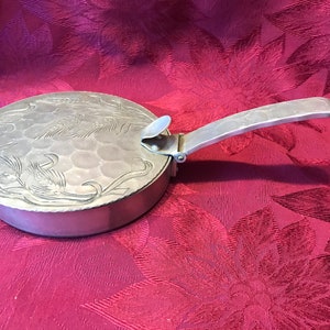 May include: A silver, round, embossed metal container with a hinged handle. The container has a floral design and a textured surface.
