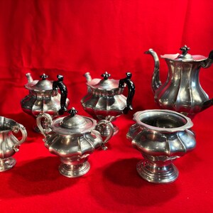 May include: A silver tea set comprising a teapot, sugar bowl, creamer, and three small teacups. The set features a fluted design and black handles. The set is displayed on a red surface.