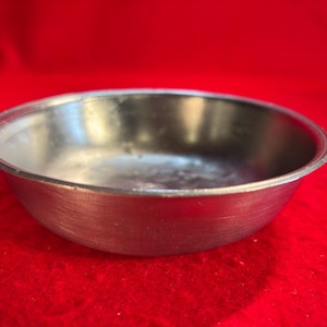 May include: A small, round, metallic bowl with a smooth, reflective interior surface and a brushed exterior finish. The bowl features a simple, curved rim and a shallow basin, presenting a clean, utilitarian design with a neutral silver-toned appearance.