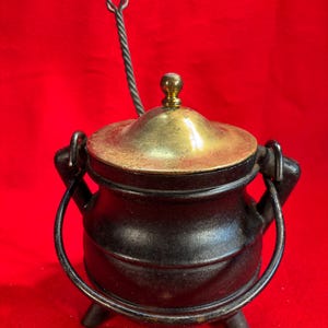 Puede incluir: Un pequeño caldero de hierro fundido negro con tapa de latón y cuchara a juego. El caldero tiene un asa a cada lado y tres patas pequeñas. La cuchara tiene un mango en forma de corazón. El fondo es rojo liso.