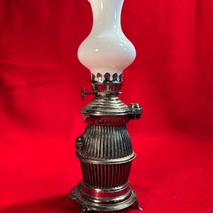 May include: A vintage oil lamp with a white glass shade and a silver-colored metal base. The lamp has a decorative design with vertical lines on the base and a small handle for adjusting the flame. The lamp is a classic decorative item.