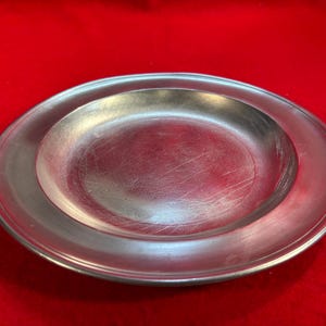 May include: A silver-coloured, oval-shaped plate with a slightly raised rim. The plate has a smooth, reflective surface with visible scratches. The background is a solid red colour. This plate is suitable for serving food.