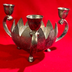 May include: Silver candle holder with three candle cups, shaped like a lotus flower with a scalloped edge. The holder has three legs and a heart-shaped handle on each side.