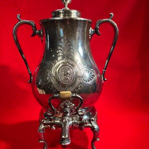 May include: A silver-toned metal beverage dispenser featuring a central medallion with a profile portrait, two curved side handles, a lid with a finial, and a spigot with a small wooden accent. The vessel stands on four ornate, claw-footed legs.