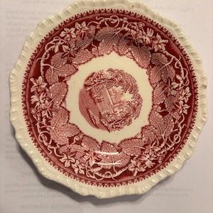 May include: A round ceramic saucer with a scalloped edge, featuring a deep red transferware pattern on a cream background. The center depicts a small landscape scene, while the border is decorated with intricate grape leaves and vine motifs.