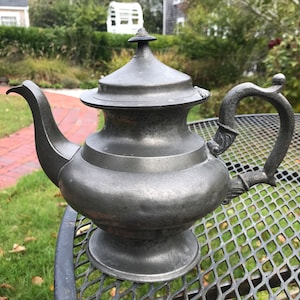May include: A vintage pewter teapot with a rounded body, a domed lid, and a curved handle. The teapot is sitting on a black metal table with a grid pattern.