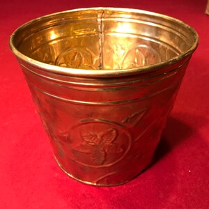 Könnte beinhalten: Ein goldfarbener Metalltopf mit einem dekorativen, geprägten Muster aus Blättern. Der Topf ist rund und hat einen breiten Rand.