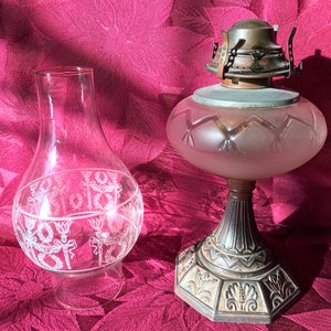May include: Two vintage oil lamp chimneys. One is clear glass with a white etched design. The other is frosted glass with a geometric design. Both are on a red fabric background.