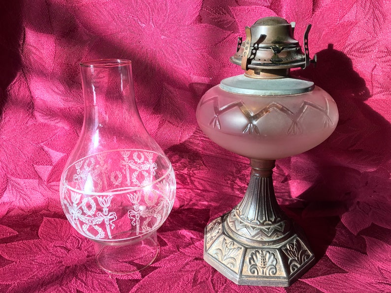 May include: Two vintage oil lamp chimneys. One is clear glass with a white etched design. The other is frosted glass with a geometric design. Both are on a red fabric background.
