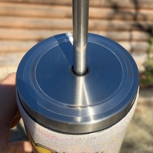 May include: A stainless steel tumbler with a lid and straw. The tumbler has a white glitter finish and a sunflower design with the words "Be Sunshine". The straw is inserted into the lid.
