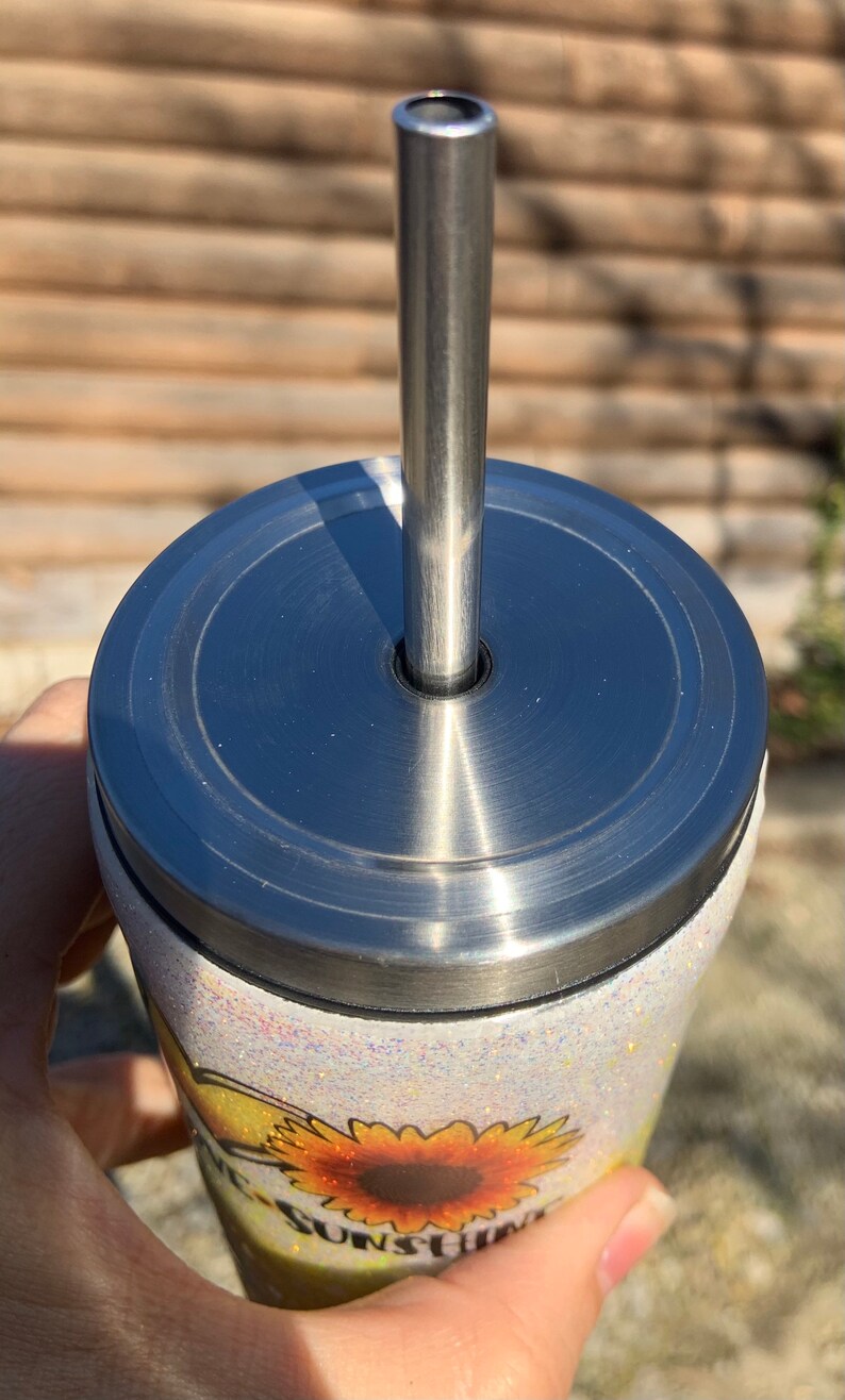 May include: A stainless steel tumbler with a lid and straw. The tumbler has a white glitter finish and a sunflower design with the words "Be Sunshine". The straw is inserted into the lid.