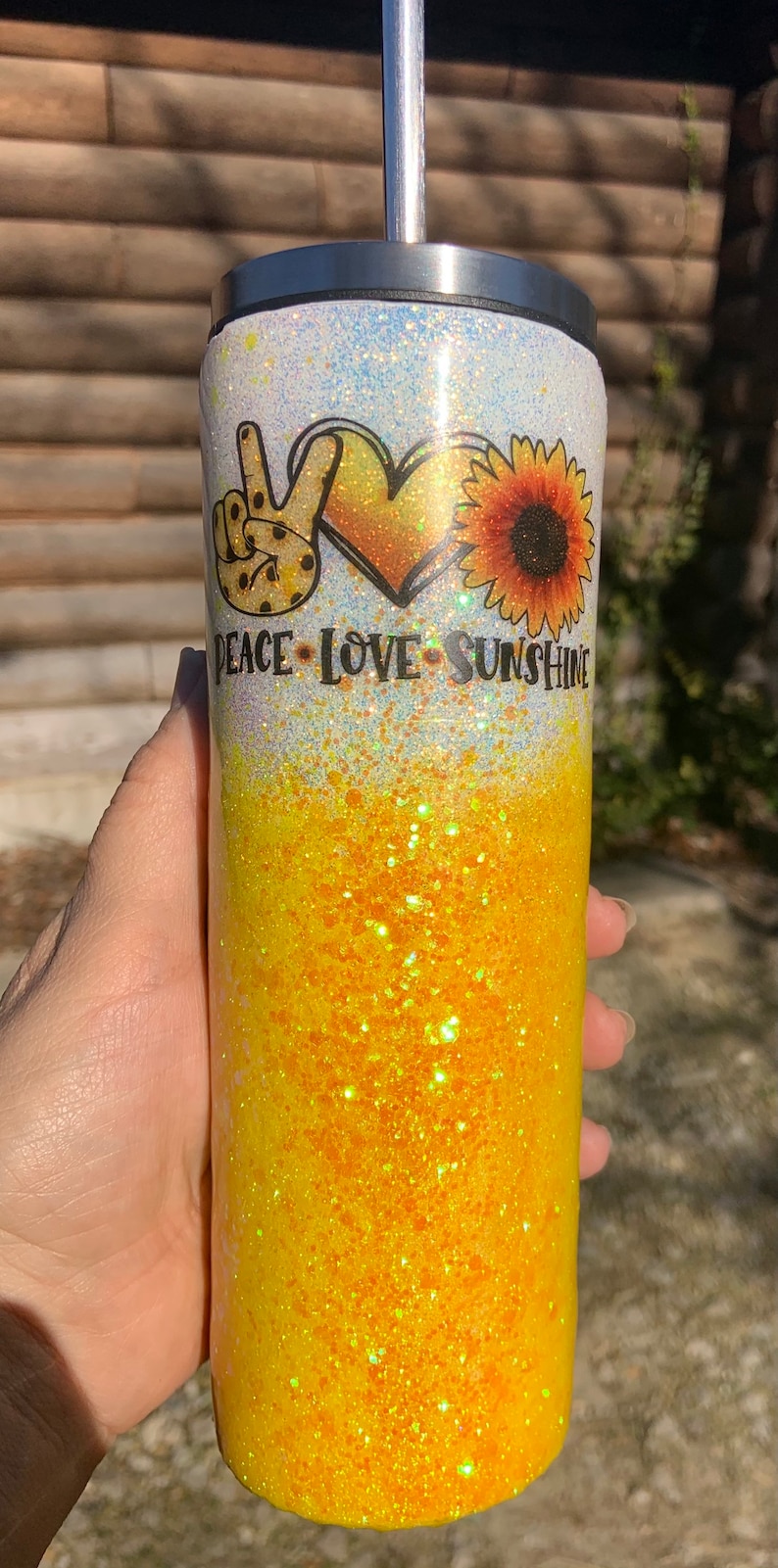 May include: Glitter-covered tumbler with a peace sign, heart, and sunflower design. The tumbler has a gradient of yellow and white glitter with the words "PEACE LOVE SUNSHINE". A stainless steel straw is inserted in the lid.