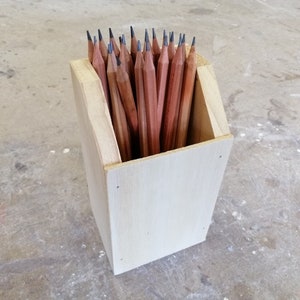 May include: A wooden pencil holder with a light brown finish. The holder is filled with a variety of pencils, all with black graphite tips.
