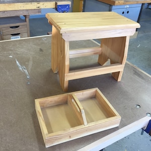 May include: A small, unfinished wooden step stool and a matching tray with a handle. The stool has a rectangular top and a simple, sturdy design. The tray is divided into two sections. Both items are made of light-coloured wood.