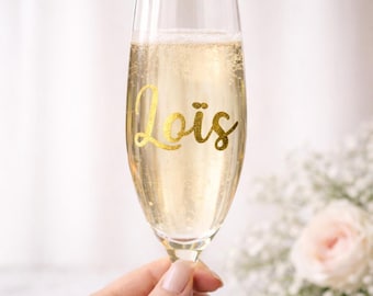 May include: A champagne flute filled with bubbly liquid, featuring the name "Lois" in gold script. The glass is held in a hand, with a soft-focus background of flowers.
