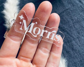 May include: A clear acrylic keychain in a cloud shape with the name "Morris" in white script. The keychain has a silver metal ring and is held in a hand. The background is a dark blue fabric.
