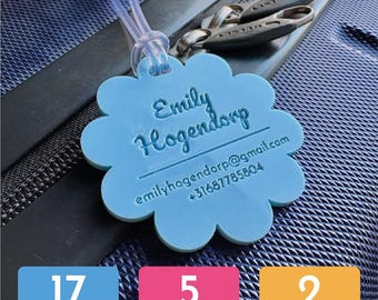 May include: A light blue luggage tag with a scalloped edge, featuring the name "Emily Hogendorp", email, and phone number. The tag is attached to a suitcase with a clear plastic loop. The image also shows the text "17 kleuren", "5 vormen", and "2 lettertypes".