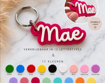 May include: A custom keychain featuring the name Mae in white script lettering layered over a hot pink scalloped base. The keychain includes a silver metal ring. The image displays text indicating availability in 12 fonts and 17 color options.