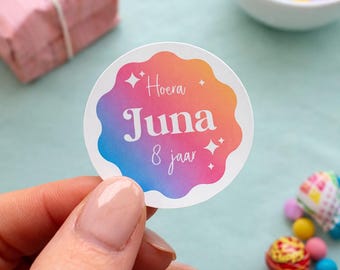 May include: A round sticker with a colorful gradient design, featuring the text "Hoera Juna 8 jaar". The sticker has a scalloped edge and is held by a person. Other items in the image include a gift box, candies, and lollipops.