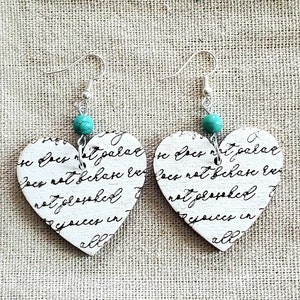 May include: A pair of heart-shaped earrings with a turquoise bead on each earring. The hearts are made of wood and have a white background with black script writing. The script reads "Docs not palar in for not palar in Docs not fchase Exa not josked joices in all nataske rejoices in all".