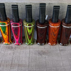 May include: Eight duck calls arranged in a row, each featuring a black plastic mouthpiece and a cylindrical wooden body with a distinct color pattern, including brown, green, pink, lime, dark brown, orange, and red, all marked with the letters CR.