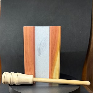 May include: A wooden box with a silver stripe down the center, flanked by reddish-brown and light-colored wood panels. A small, light-colored wooden mallet rests in front of the box, suggesting a craft or woodworking theme.