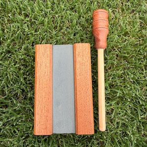 May include: A rectangular sharpening stone set between two parallel wooden blocks, accompanied by a separate wooden tool featuring a cylindrical handle with three horizontal grooves and a long, slender, light-colored wooden shaft.
