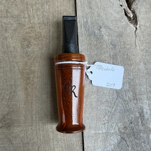 May include: A wooden duck call with a dark brown finish and a black mouthpiece. The call has the initials "GR" engraved on the side and a tag that reads "Cocobolo" and "209". The call is made of cocobolo wood.