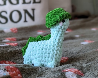 May include: A crocheted green dinosaur toy with a green hat, sitting on a green blanket with red mushroom designs.
