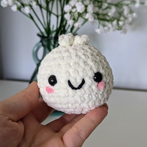 May include: A white crocheted plush toy shaped like a dumpling with a smiling face, black eyes, and pink cheeks.