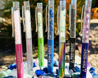May include: Six crystal-filled pens with silver caps and clear barrels. The pens are arranged in a row on a bed of colourful crystals. Each pen has a different colour and crystal type. The pens are labelled "Pen-Energy Crystal Power Pens".