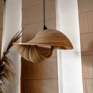 Op de afbeelding: Een unieke, handgemaakte hanglamp. De lampenkap is gemaakt van gelaagde, gebogen houten strips, waardoor een wervelend, open ontwerp ontstaat. De lamp hangt aan een zwart snoer, tegen een neutrale achtergrond.