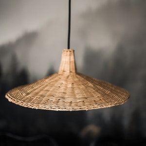 K&ouml;nnte beinhalten: Eine geflochtene H&auml;ngeleuchte aus Weide mit einem breiten, flachen, kegelf&ouml;rmigen Schirm, der an einem schwarzen Kabel h&auml;ngt.