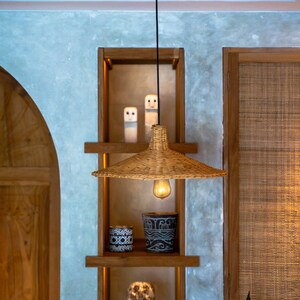 K&ouml;nnte beinhalten: Eine geflochtene H&auml;ngeleuchte aus Weide mit einer Gl&uuml;hbirne, die an einem schwarzen Kabel in einem Holzregalsystem mit zwei kleinen wei&szlig;en Keramikfiguren, zwei dekorativen T&ouml;pfen und einer Holzmaske h&auml;ngt.