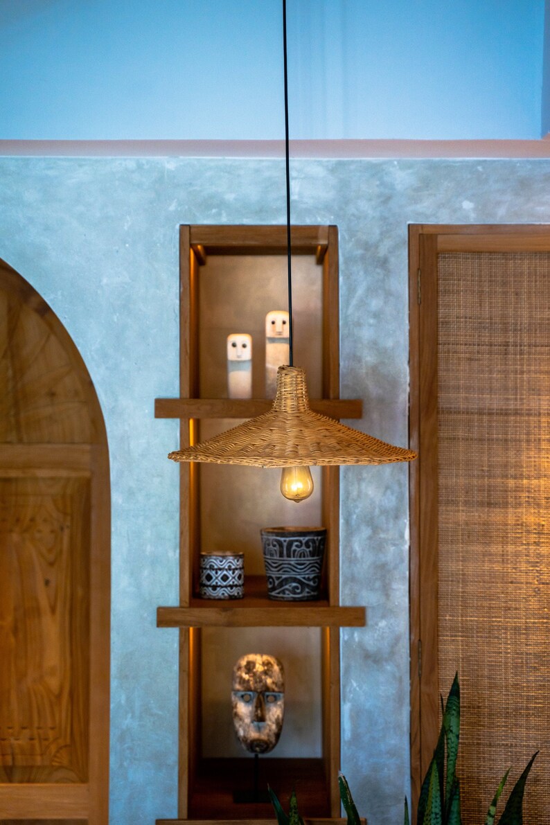 K&ouml;nnte beinhalten: Eine geflochtene H&auml;ngeleuchte aus Weide mit einer Gl&uuml;hbirne, die an einem schwarzen Kabel in einem Holzregalsystem mit zwei kleinen wei&szlig;en Keramikfiguren, zwei dekorativen T&ouml;pfen und einer Holzmaske h&auml;ngt.