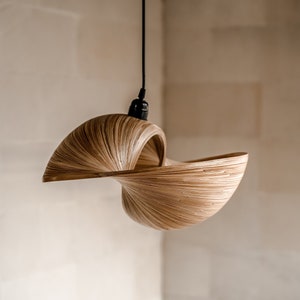 Op de afbeelding: Een unieke hanglamp met een wervelend, open ontwerp. De lamp is gemaakt van dunne, lichtbruine houten strips, waardoor een getextureerd, gelaagd effect ontstaat. Hij hangt aan een zwart snoer en is ideaal voor een natuurlijke uitstraling.