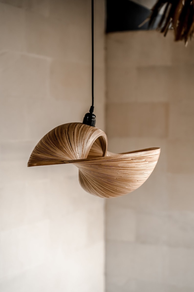 Op de afbeelding: Een unieke hanglamp met een wervelend, open ontwerp. De lamp is gemaakt van dunne, lichtbruine houten strips, waardoor een getextureerd, gelaagd effect ontstaat. Hij hangt aan een zwart snoer en is ideaal voor een natuurlijke uitstraling.
