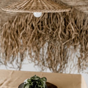 K&ouml;nnte beinhalten: Eine geflochtene H&auml;ngeleuchte aus Weide h&auml;ngt &uuml;ber einem Holztablett mit einem gr&uuml;nen, bl&auml;ttrigen Salat auf einer beigefarbenen Tischdecke. Der Hintergrund ist eine Wand mit einem gewebten Wandbehang in Braun und Beige.