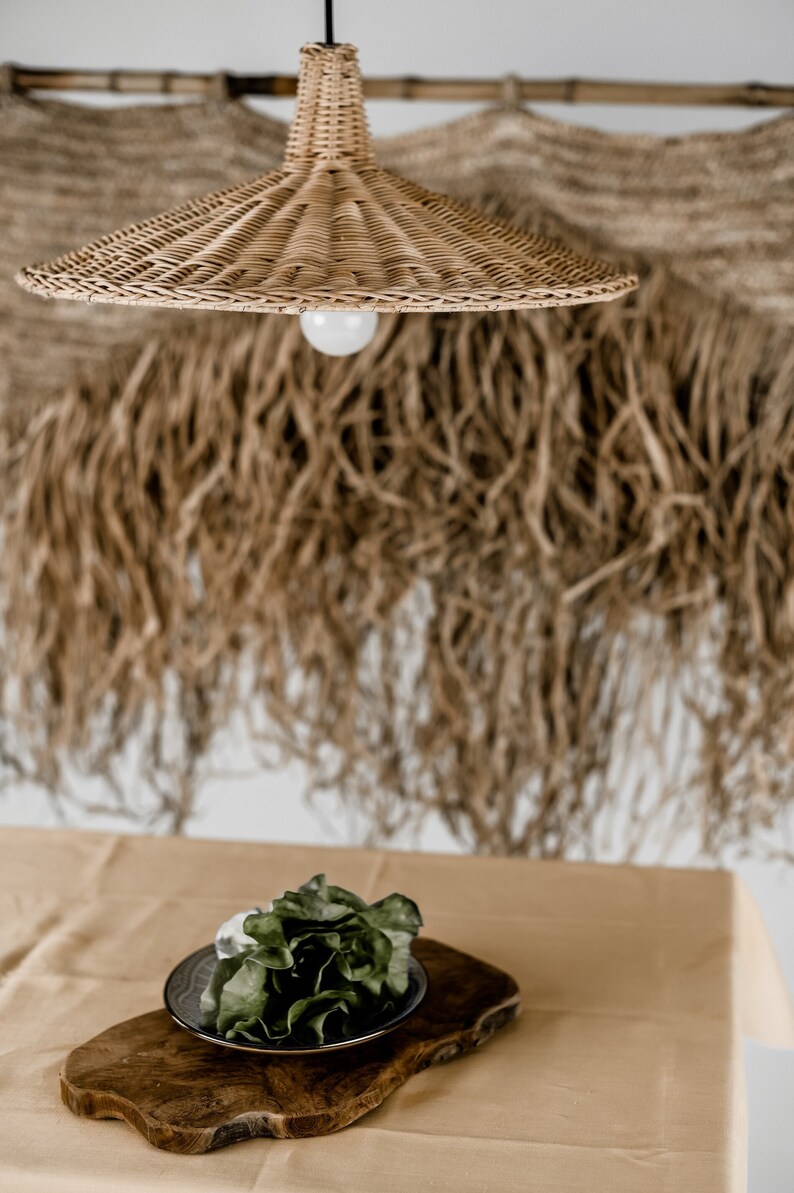 K&ouml;nnte beinhalten: Eine geflochtene H&auml;ngeleuchte aus Weide h&auml;ngt &uuml;ber einem Holztablett mit einem gr&uuml;nen, bl&auml;ttrigen Salat auf einer beigefarbenen Tischdecke. Der Hintergrund ist eine Wand mit einem gewebten Wandbehang in Braun und Beige.