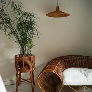 K&ouml;nnte beinhalten: Ein Sofa aus Weidengeflecht mit einem wei&szlig;en Kissen steht in einem Raum mit einer Topfpflanze und einer geflochtenen Pendelleuchte.