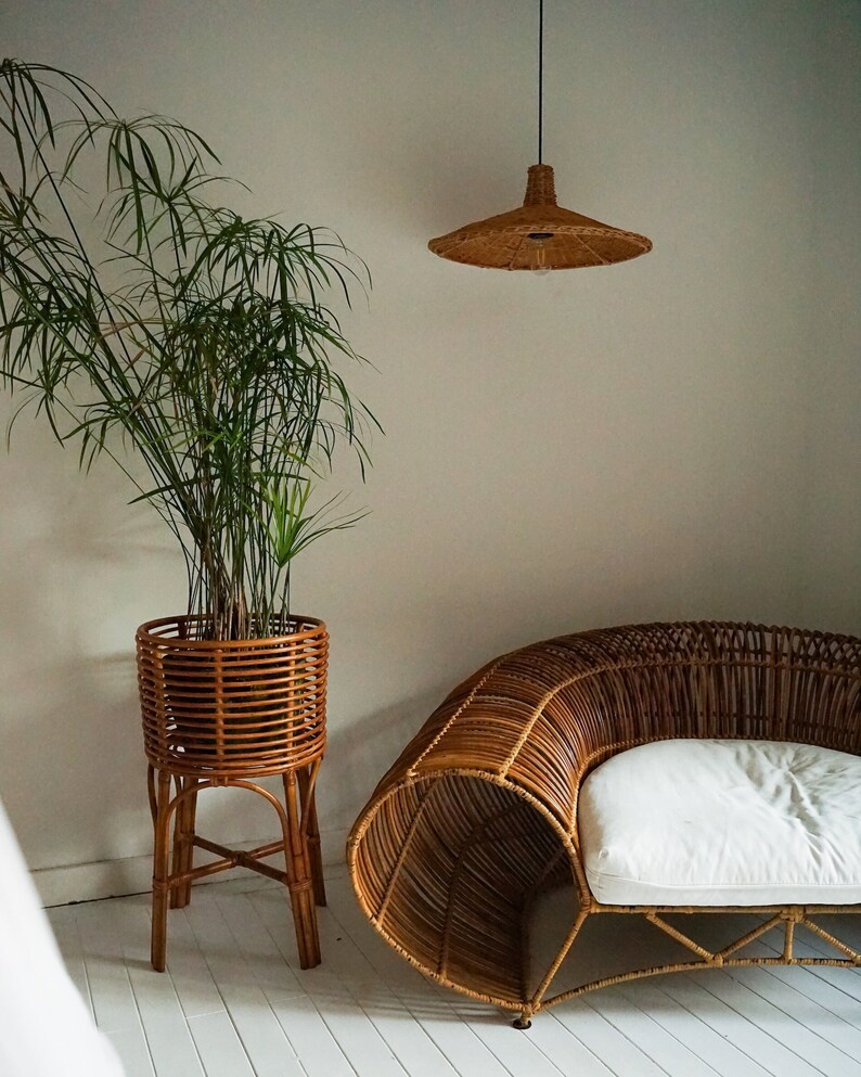 K&ouml;nnte beinhalten: Ein Sofa aus Weidengeflecht mit einem wei&szlig;en Kissen steht in einem Raum mit einer Topfpflanze und einer geflochtenen Pendelleuchte.