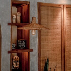 K&ouml;nnte beinhalten: Eine geflochtene H&auml;ngeleuchte aus Weide mit einer warmwei&szlig;en Gl&uuml;hbirne, die an einem schwarzen Kabel vor einem Holzregal mit Dekorationsgegenst&auml;nden h&auml;ngt. Das Regal ist Teil einer gr&ouml;&szlig;eren Holzkonstruktion mit einer geflochtenen Holzt&uuml;r dahinter.