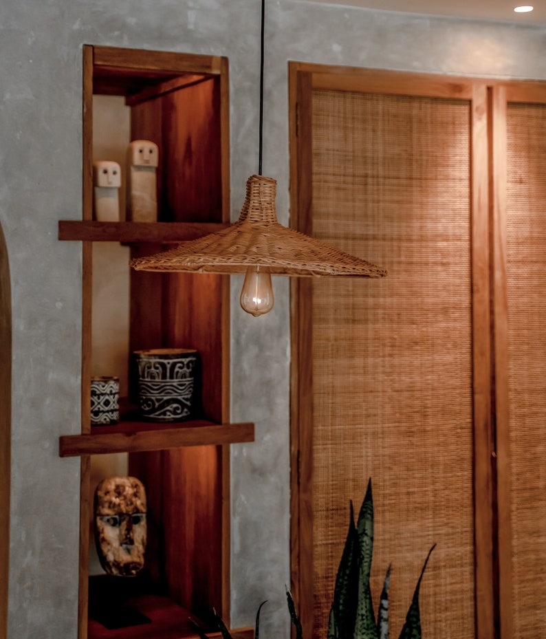 K&ouml;nnte beinhalten: Eine geflochtene H&auml;ngeleuchte aus Weide mit einer warmwei&szlig;en Gl&uuml;hbirne, die an einem schwarzen Kabel vor einem Holzregal mit Dekorationsgegenst&auml;nden h&auml;ngt. Das Regal ist Teil einer gr&ouml;&szlig;eren Holzkonstruktion mit einer geflochtenen Holzt&uuml;r dahinter.