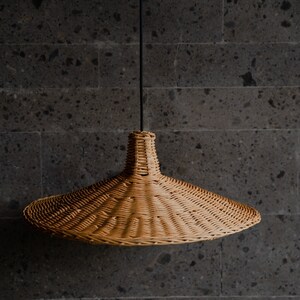 K&ouml;nnte beinhalten: Eine geflochtene H&auml;ngeleuchte aus Weide h&auml;ngt an einem schwarzen Kabel vor einer dunkelgrauen, strukturierten Wand.
