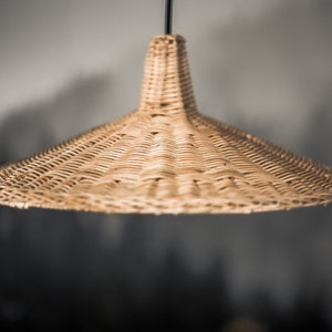 K&ouml;nnte beinhalten: Eine geflochtene H&auml;ngeleuchte aus Weide mit einem breiten, konischen Schirm. Die Leuchte h&auml;ngt an einem schwarzen Kabel.