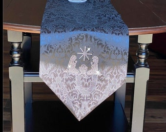 May include: A black table runner with a silver damask pattern and a rhinestone nativity scene design. The runner is on a dark wood table with a clear glass vase.